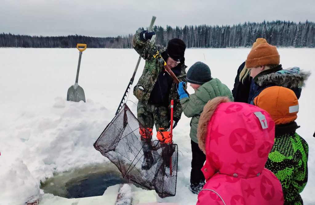 Mies esittelee lapsiryhmälle kalastusvälineitä avannon äärellä.